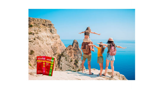 A vibrant outdoor scene featuring assorted grass-fed beef snack sticks, including Original Beef and Hatch Green Chile, surrounded by lush greenery and hints of a hiking trail under a clear blue sky.