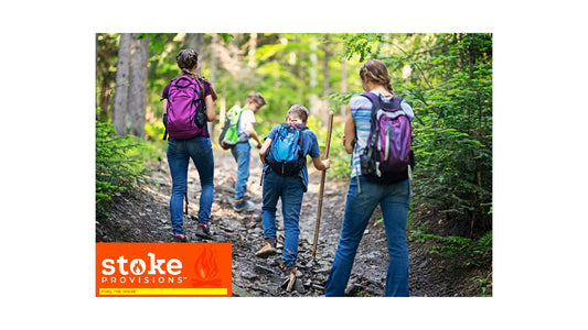 A diverse group of young adults hiking and enjoying a snack break with beef snack sticks in a lush, green outdoor setting under a clear sky.